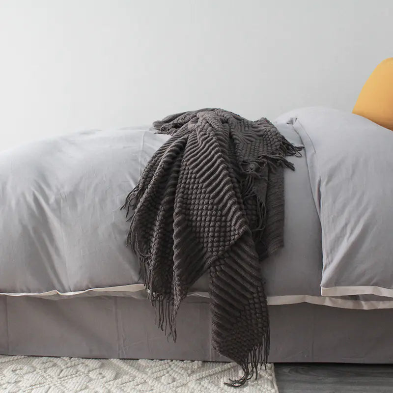 Gray bed with a dark gray knitted throw blanket and a yellow pillow.