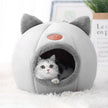 Cat peeking out from a gray and white pet bed with cat ears on a white surface.