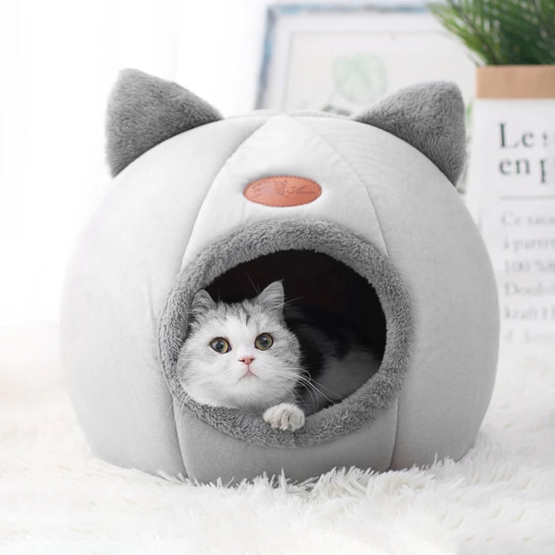 Cat peeking out from a gray and white pet bed with cat ears on a white surface.