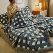 Person sitting on a bed with a blue and white checkered blanket, holding a cup.
