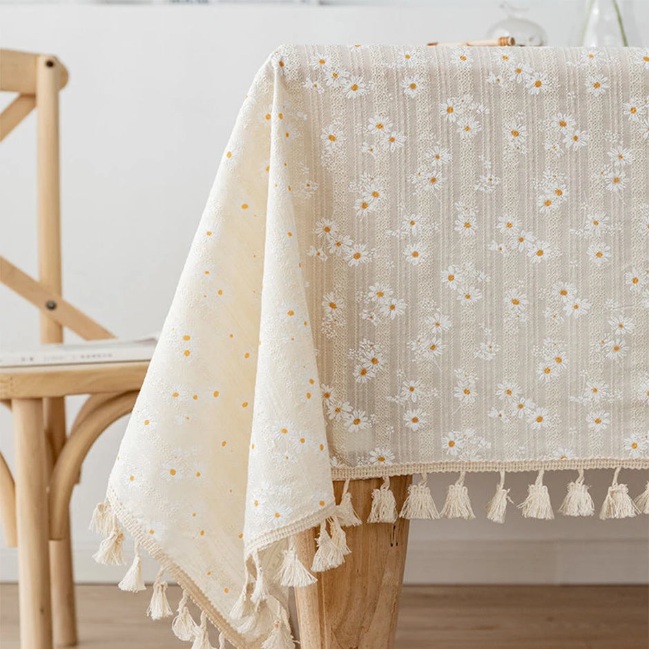 Beige floral tablecloth with tassels on a wooden table and chair.