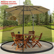 Beige patio umbrella with mosquito net over a wooden outdoor table and chairs on a grassy area.