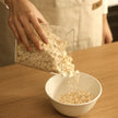 Person pouring oats from a transparent container into a white bowl on a wooden table.