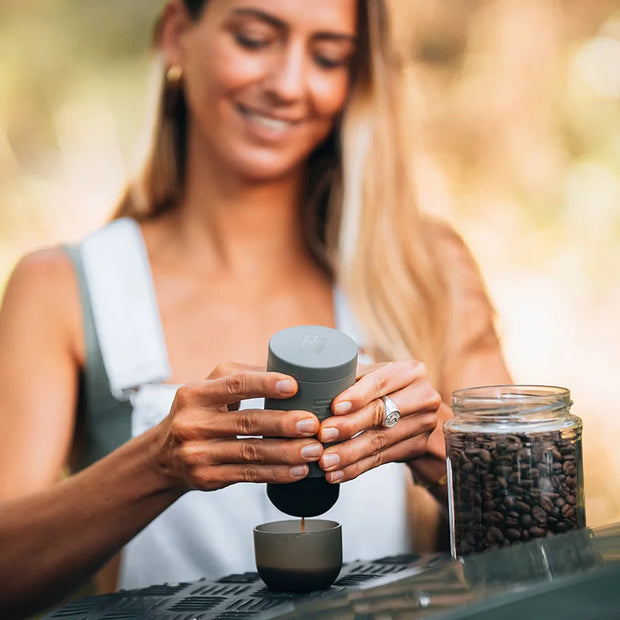 Woman using a portable coffee maker outdoors with a jar of coffee beans nearby.