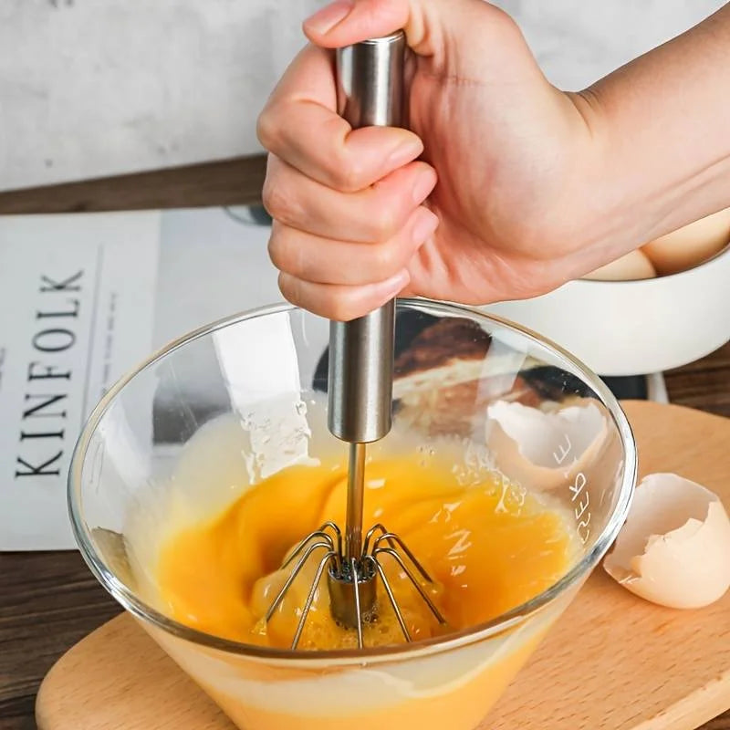 Hand using a manual egg beater in a glass bowl with eggs.