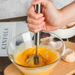 Hand using a manual egg beater in a glass bowl with eggs.