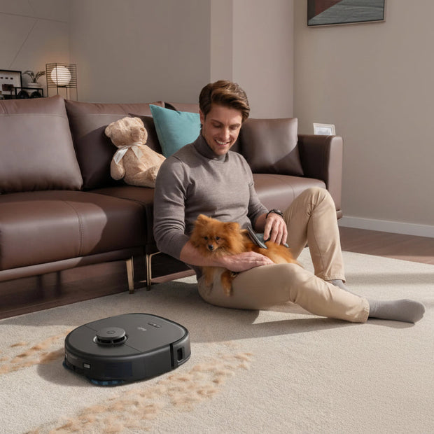 Person sitting on the floor with a dog next to a robot vacuum cleaner in a living room.
