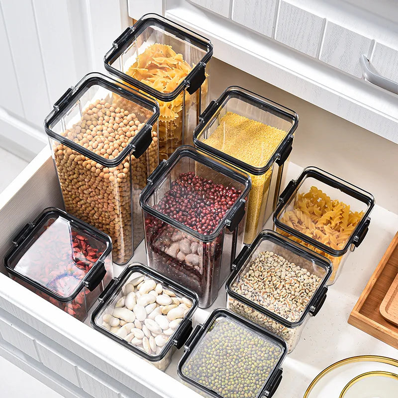 Set of clear food storage containers with black lids containing various dry goods in a kitchen setting.