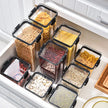 Set of clear food storage containers with black lids containing various dry goods in a kitchen setting.