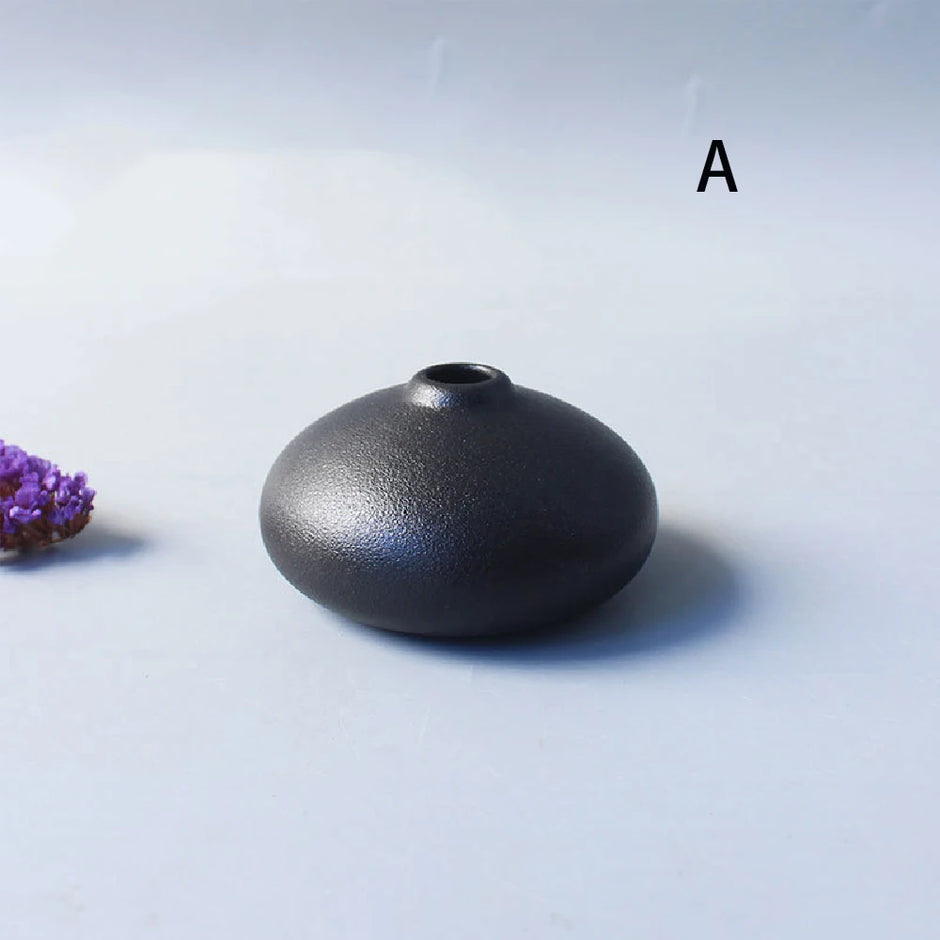 Black ceramic vase on a light gray background with a purple flower to the left.