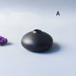 Black ceramic vase on a light gray background with a purple flower to the left.