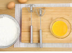 Two metal beaters on a wooden cutting board with eggs, flour, and a bowl of yellow mixture.