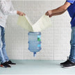 Two people pouring water from a large blue container into a smaller green container against a white brick wall.