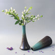 Two ceramic vases with a branch of white flowers and green leaves on a light gray background.