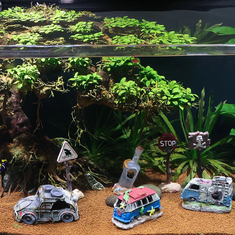 Decorative aquarium scene with miniature cars, bottles, and signs on a sandy bottom with green plants.