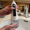 Hand holding a waterpulse dental device over a sink with bathroom fixtures in the background