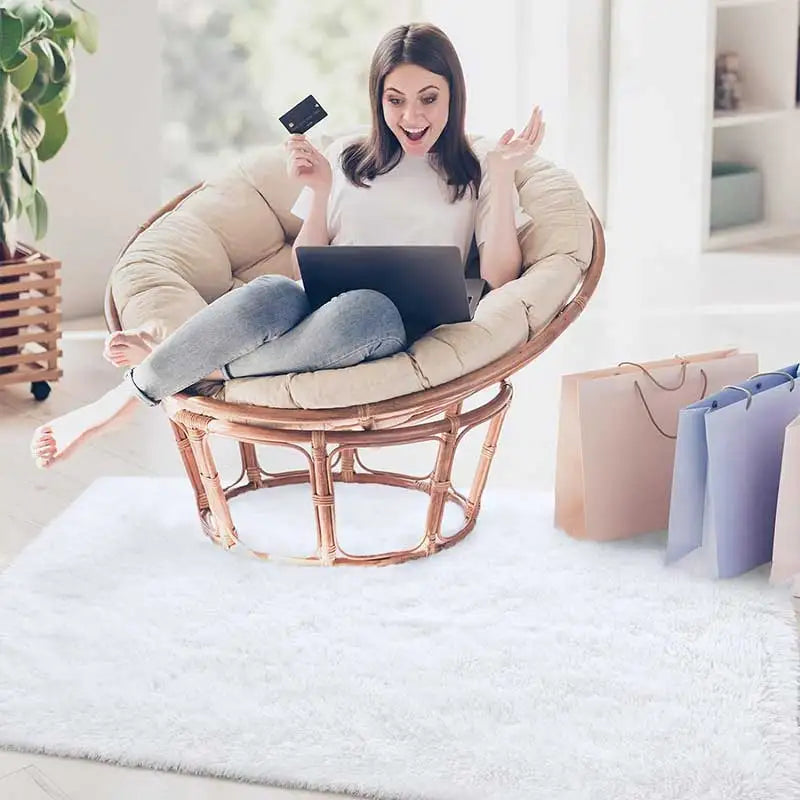 Woman sitting on a round rattan chair with a laptop and credit card, surrounded by shopping bags in a bright room.