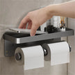 Bathroom shelf with paper towels, phone, and other items on a tiled wall.
