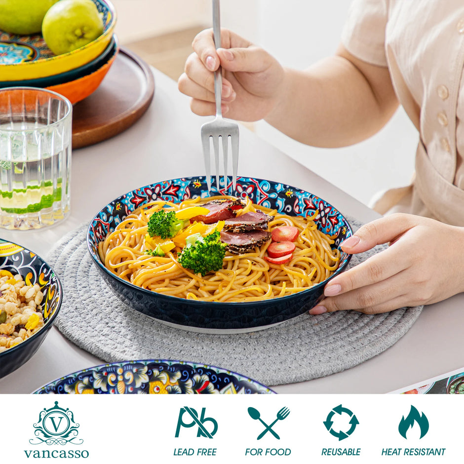 Person holding a bowl of spaghetti with colorful plates and glasses on a table, featuring the Vancasso brand.
