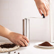 Person using a manual coffee grinder with coffee beans and ground coffee on a white surface.