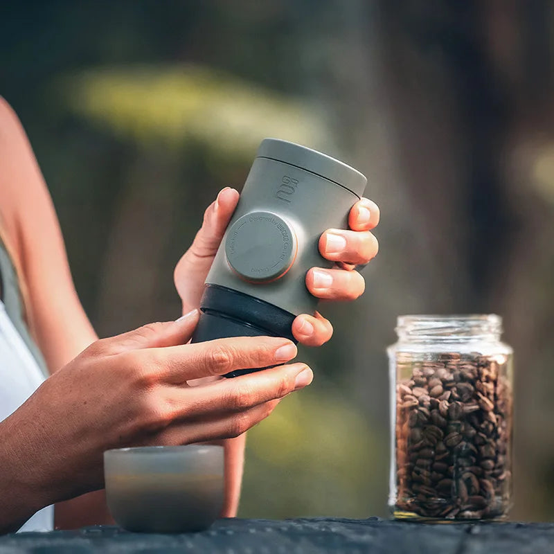 Person holding a gray travel mug with a blurred background of coffee beans and greenery