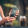 Person holding a gray travel mug with a blurred background of coffee beans and greenery