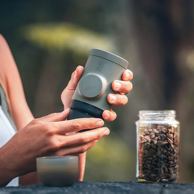 Person holding a gray travel mug with a blurred background of coffee beans and greenery