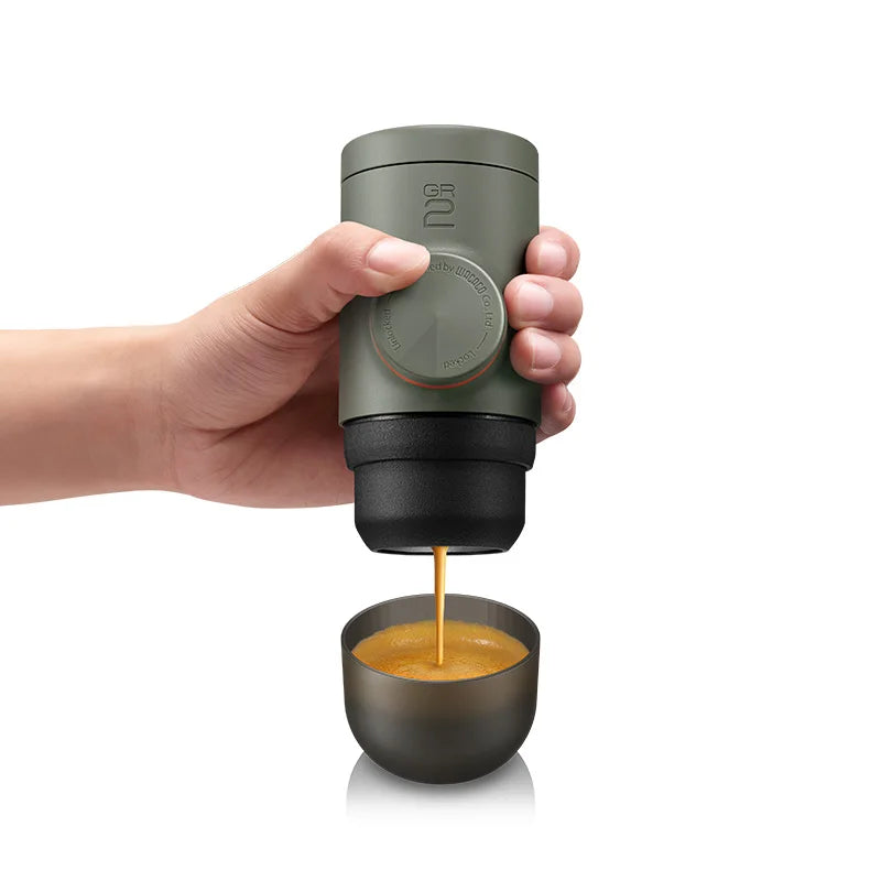 Hand holding a green portable coffee maker pouring coffee into a small cup on a white background