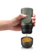 Hand holding a green portable coffee maker pouring coffee into a small cup on a white background