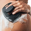 Person using a black skin exfoliating tool on their shoulder with soap suds.