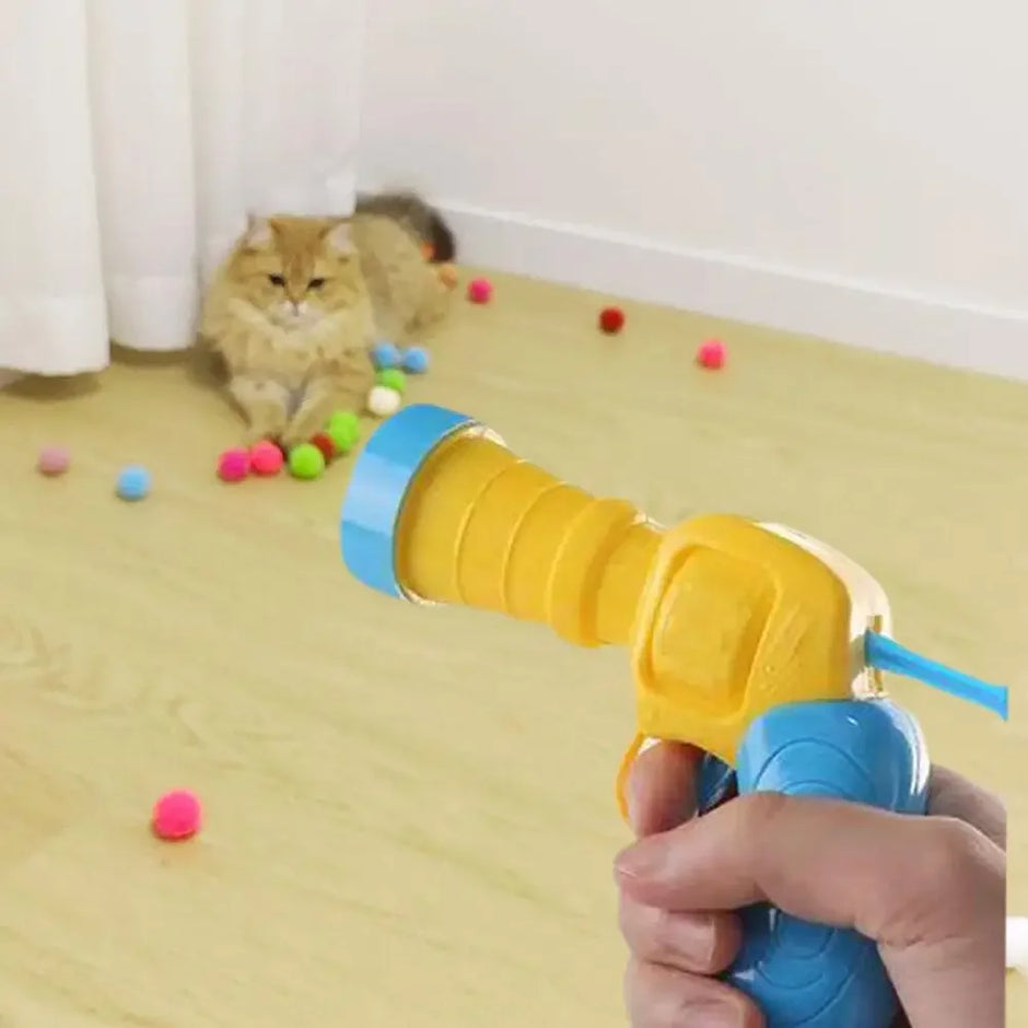 Person holding a yellow and blue toy gun with a cat playing with colorful balls on the floor.
