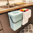 Kitchen compost bin with a lid and another bin with colorful vegetables on a kitchen counter.