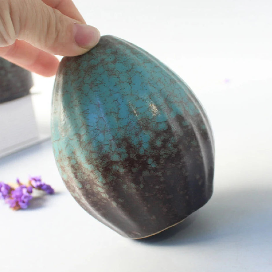 Hand holding a textured ceramic object with a blue and brown glaze on a light background.