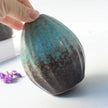 Hand holding a textured ceramic object with a blue and brown glaze on a light background.