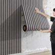 Person holding a large roll of black and gray striped wallpaper against a textured wall.