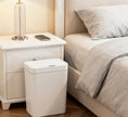 White trash bin next to a bed and nightstand in a bedroom setting