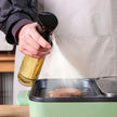 Person using a bottle of oil to cook a steak on a grill.