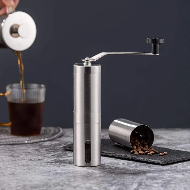 Handheld coffee grinder with coffee beans on a dark surface, with a blurred background of a person pouring coffee.