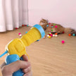Person using a toy gun to shoot colorful balls at a cat on a wooden floor.