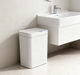 White trash bin next to a bathroom sink with a light gray wall and floor.