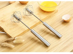 Two metal whisk attachments on a wooden surface with eggs and a bowl in the background.