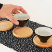 Person holding a small black cup with wooden coaster on a patterned tablecloth