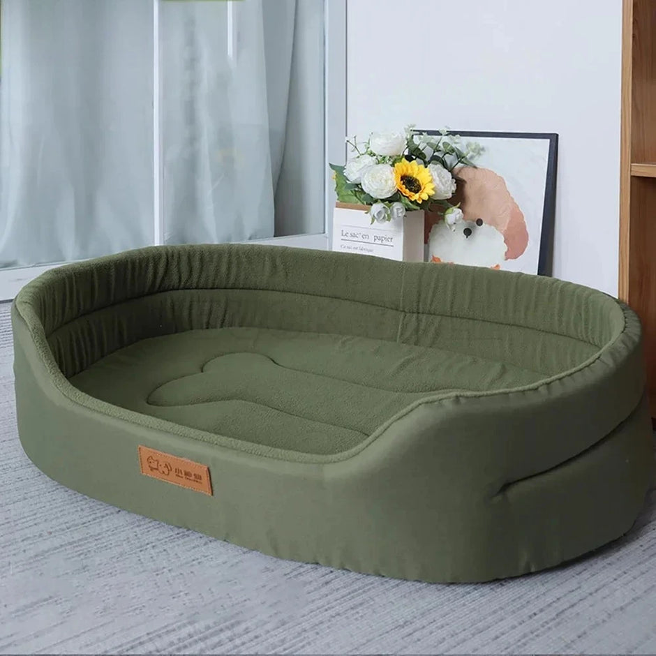 Green pet bed on a carpeted floor with a light gray curtain and floral arrangement in the background.