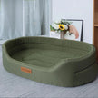 Green pet bed on a carpeted floor with a light gray curtain and floral arrangement in the background.