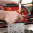 Collapsible red and gray colander being used to wash vegetables under running water.