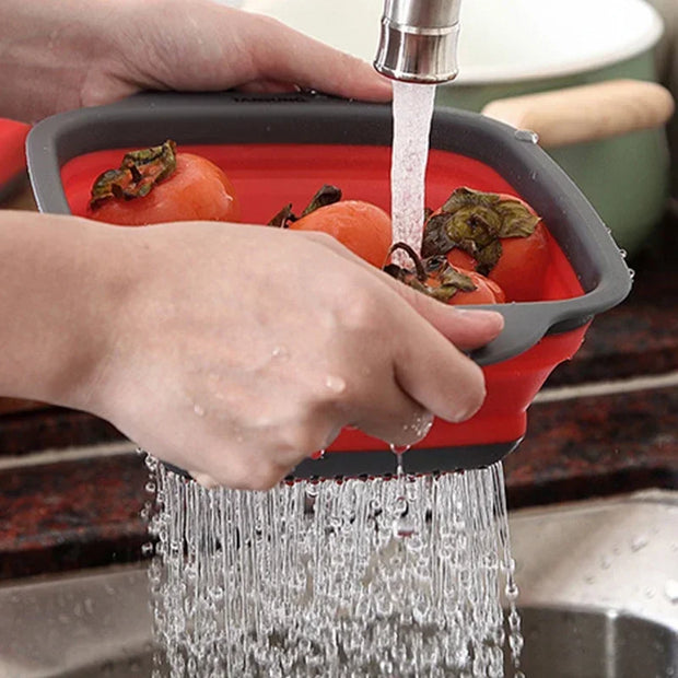 Collapsible red and gray colander being used to wash vegetables under running water.