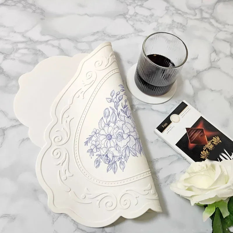 Decorative white lace doily with floral pattern on a marble surface, accompanied by a glass of dark liquid and a box of chocolates.