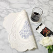 Decorative white lace doily with floral pattern on a marble surface, accompanied by a glass of dark liquid and a box of chocolates.