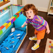 Child playing with a fish tank filled with blue sand and small fish, wearing a colorful striped shirt and orange shorts.