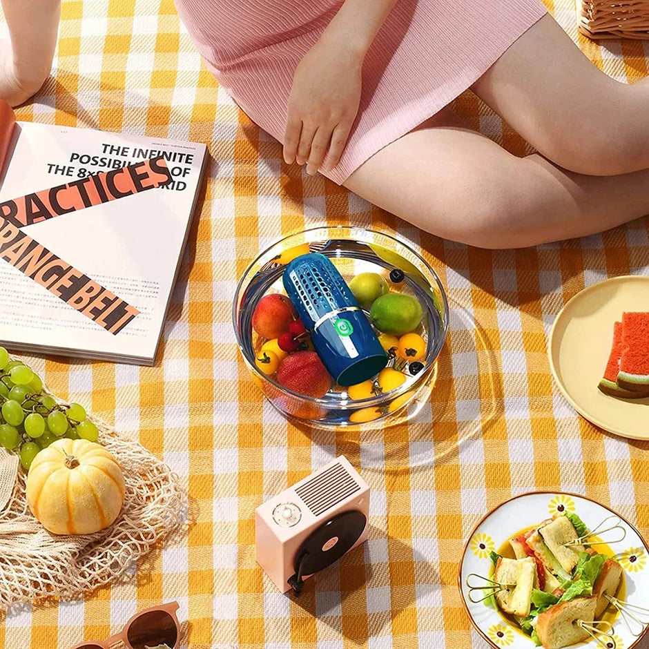 Person sitting on a yellow checkered blanket with a bowl of fruit, a book, and a small table with food.